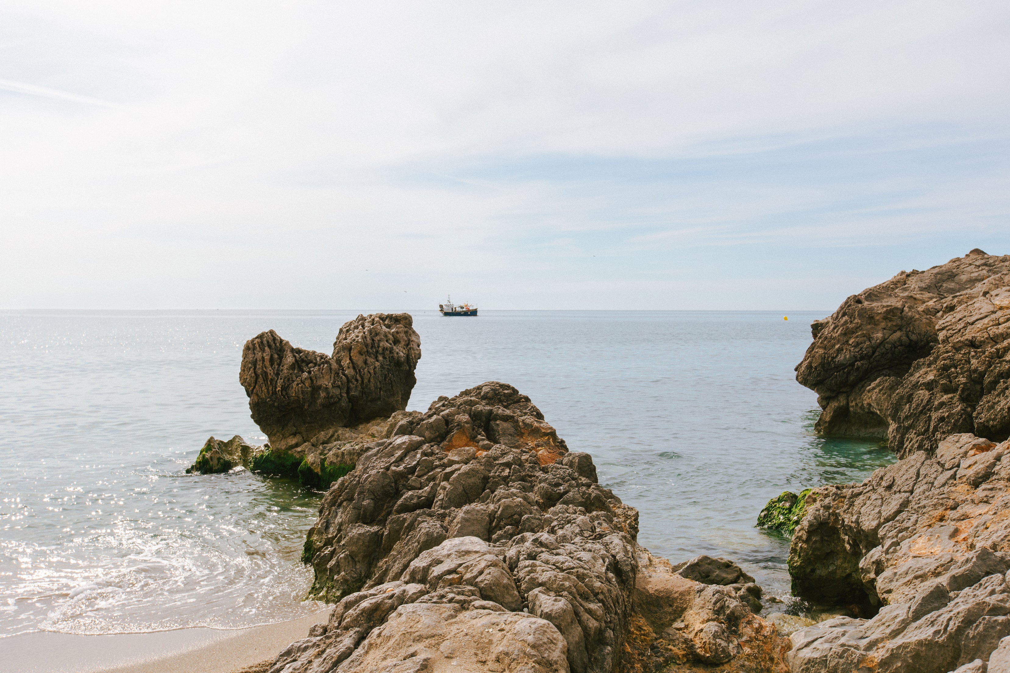 Rocky beach of Garraf Spain with fisherman boat on horizon by Bas Bains