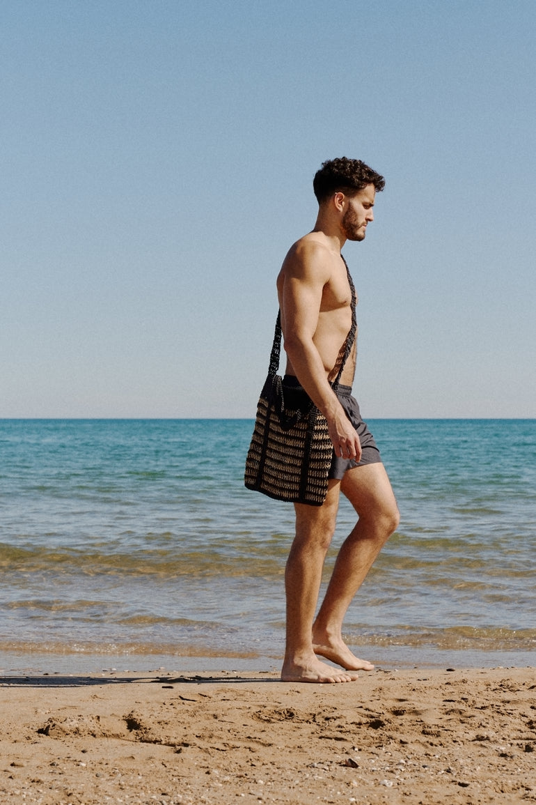 Vince Halst walking on sunny beach in black Bas Bains swimshorts wearing a Aliu Studio cross body tote bag