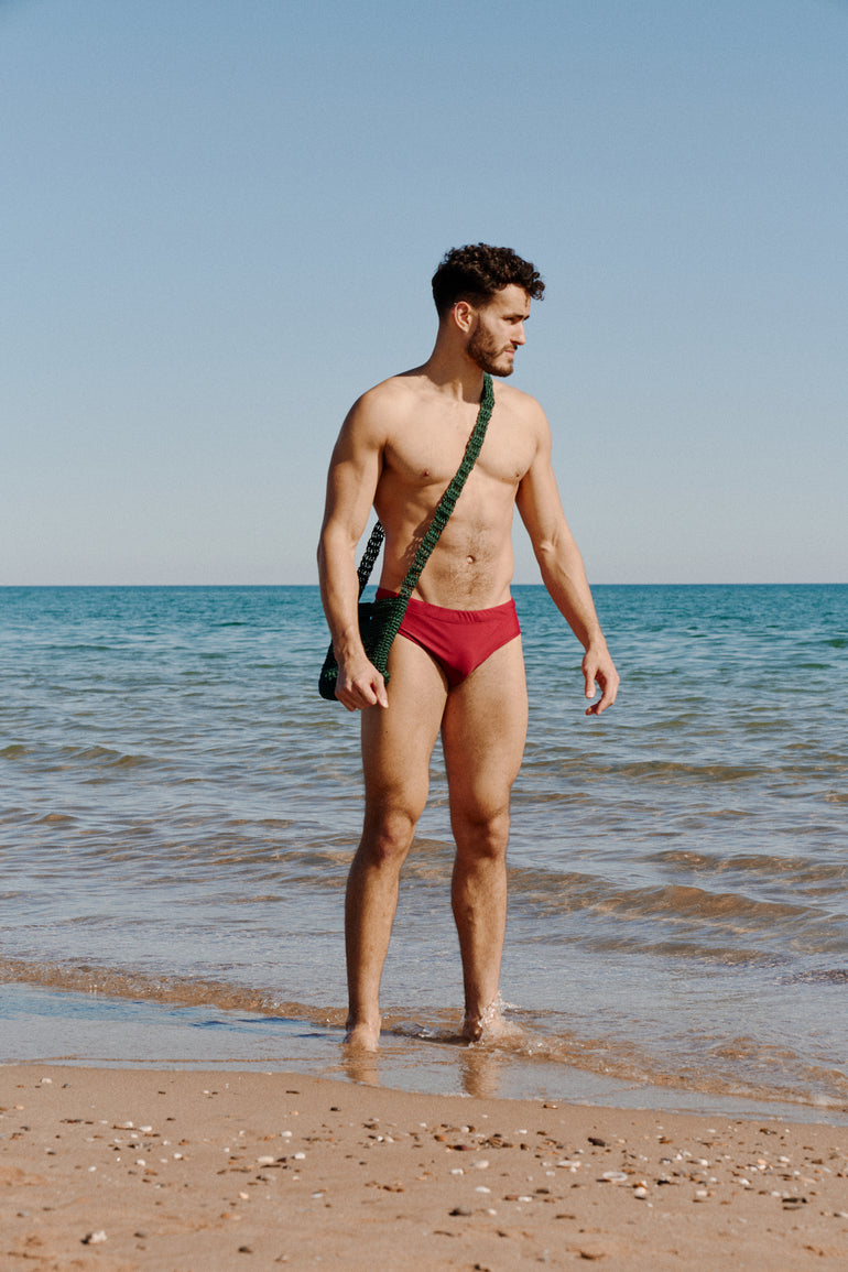 Vince Halst standing on sunny beach in black Bas Bains red slip wearing a Aliu Studio cross body bucket bag in petrol green